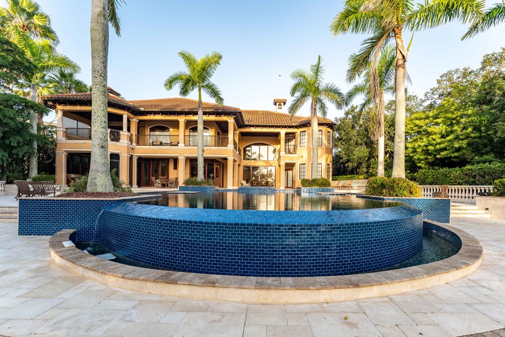 Luxury pool design with circular blue mosaic tiles, surrounded by palm trees and a spacious outdoor area, showcasing a beautiful home in a tropical setting.