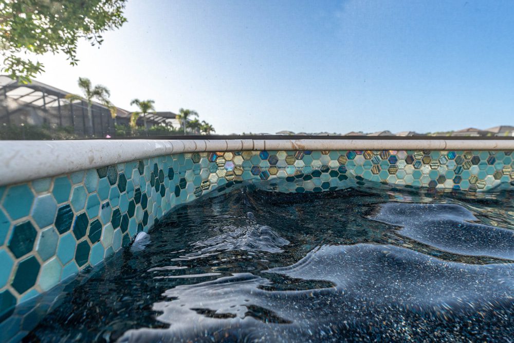 Close-up of a pool's edge featuring hexagonal mosaic tiles in shades of blue and green, with water rippling gently, set against a backdrop of palm trees and a clear sky, emphasizing Florida Family Pools' custom pool design expertise.