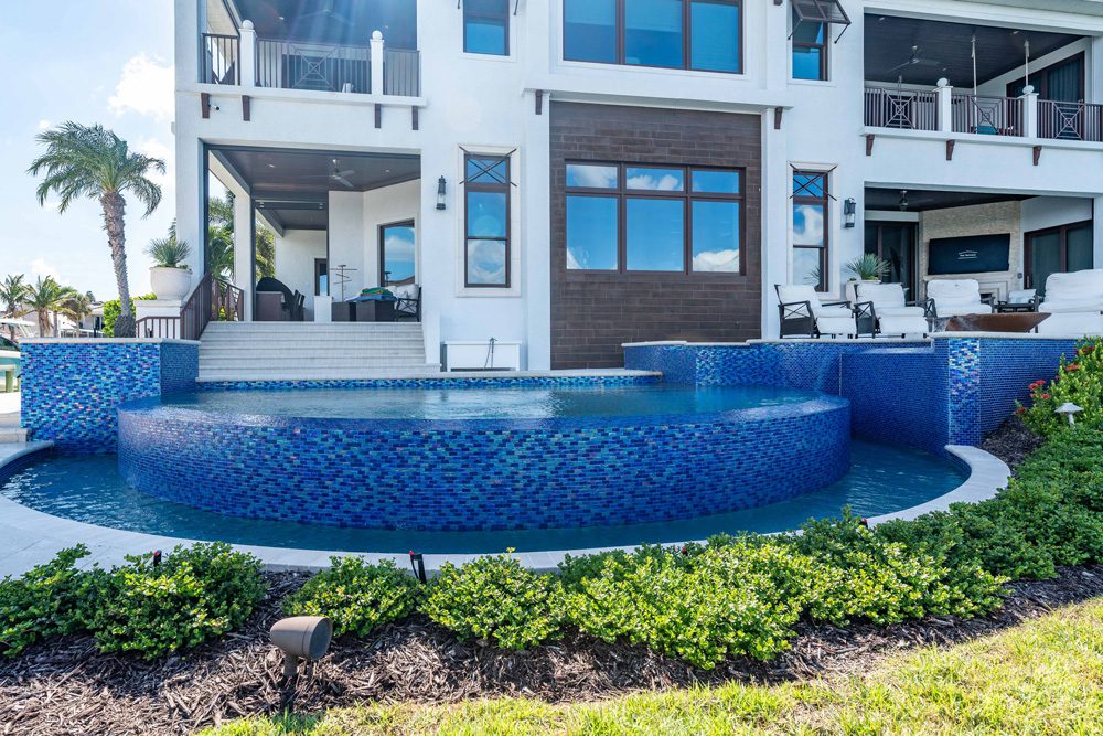Luxury custom pool with blue mosaic tiles, surrounded by lush landscaping and modern outdoor seating area, showcasing Florida Family Pools' design expertise.