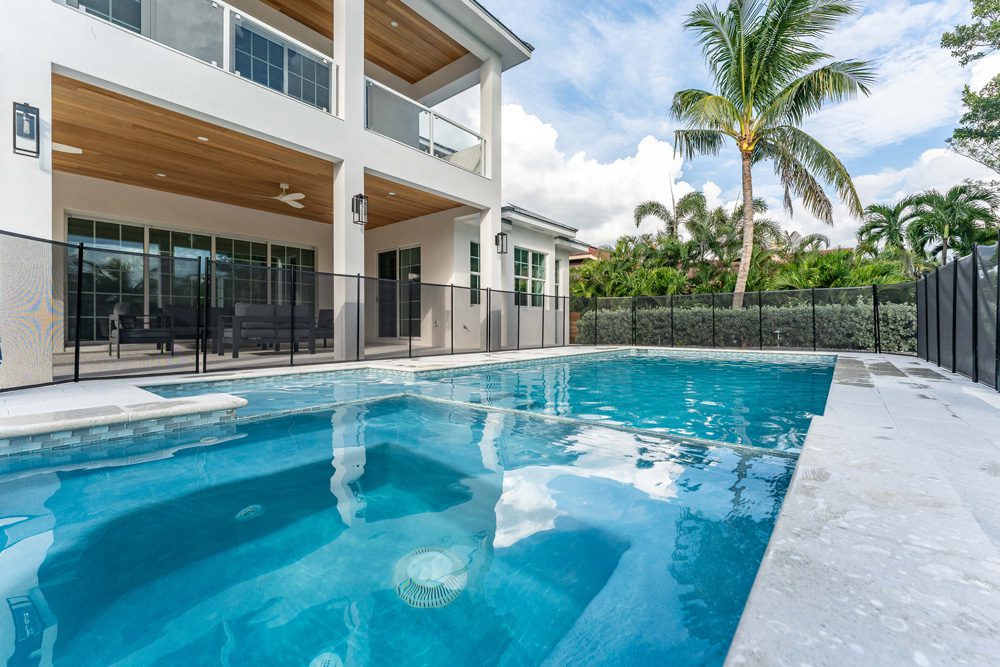Modern pool design with a clear blue water feature, surrounded by a safety fence, adjacent to a contemporary home with a spacious patio area and tropical landscaping in Sarasota, Florida.