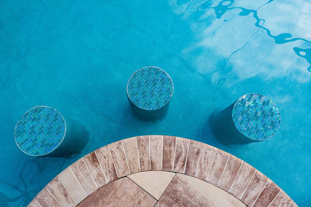 Three circular mosaic-topped stools partially submerged in a clear blue swimming pool, adjacent to a curved stone pool edge.