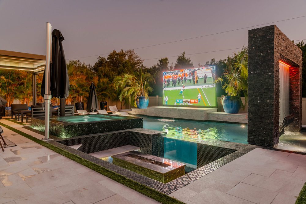 Luxury backyard pool with spa, outdoor seating, and large screen displaying a sports game, surrounded by tropical landscaping.