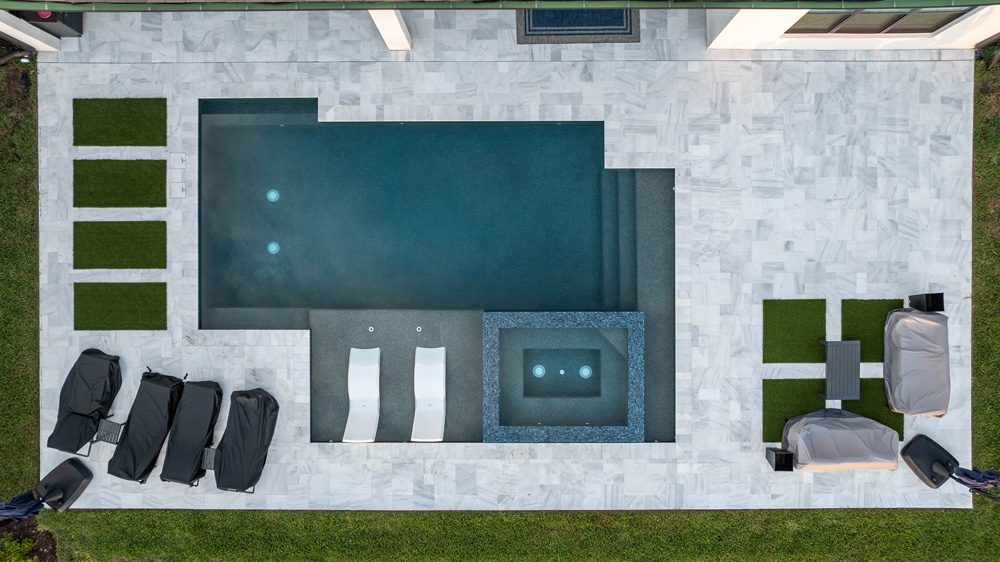 Aerial view of a modern pool design featuring a dark blue pool, spa area, and marble patio, surrounded by green grass and lounge chairs, showcasing Florida Family Pools' custom pool construction.