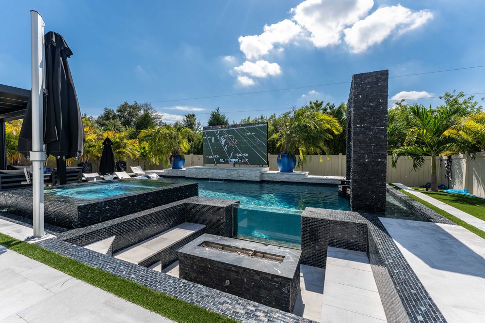 Luxury custom pool design featuring a modern layout, integrated seating, and outdoor television, surrounded by tropical landscaping and shaded by umbrellas, showcasing Florida Family Pools' expertise in pool construction.