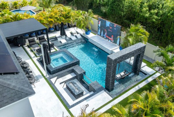 Luxurious outdoor living space featuring a modern pool, sunken fire pit, and lounge area with black umbrellas, surrounded by tropical landscaping and an outdoor TV screen, showcasing Florida Family Pools' design expertise.