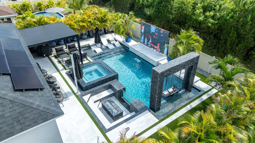 Luxury backyard pool with spa, outdoor seating, and large screen TV, surrounded by tropical landscaping, showcasing custom pool design by Florida Family Pools.