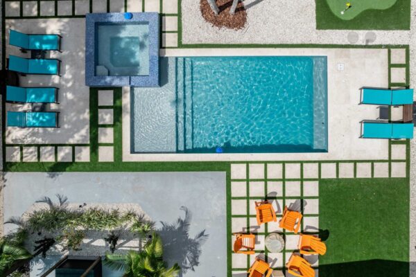 Aerial view of a resort-style outdoor space featuring a rectangular swimming pool, a spa, sun loungers, vibrant orange seating, and a putting green, surrounded by lush landscaping and geometric stone pathways, designed for enhancing guest experiences in vacation rentals.