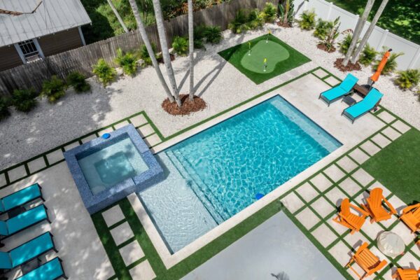 Aerial view of a resort-style outdoor space featuring a rectangular swimming pool, a spa, a putting green, and lounge chairs, surrounded by palm trees and decorative landscaping, designed for an immersive vacation experience.