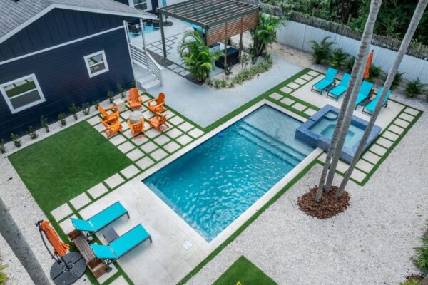 Aerial view of a resort-style outdoor space featuring a swimming pool, spa, lounge chairs, and vibrant orange seating, surrounded by palm trees and decorative landscaping, designed to enhance guest experiences in vacation rentals.
