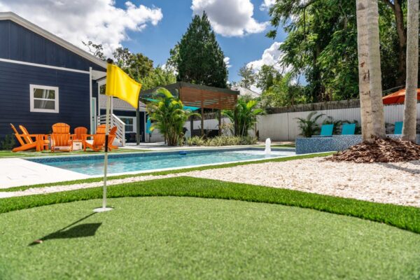 Putting green with yellow flag, swimming pool, orange lounge chairs, and tropical landscaping in a resort-style outdoor space designed for vacation rentals.
