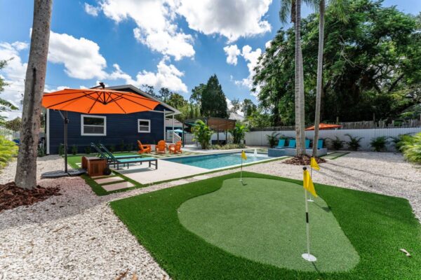 Vacation rental outdoor space featuring a putting green, swimming pool, vibrant orange umbrella, and lounge seating, surrounded by palm trees and decorative gravel, creating a resort-inspired atmosphere.