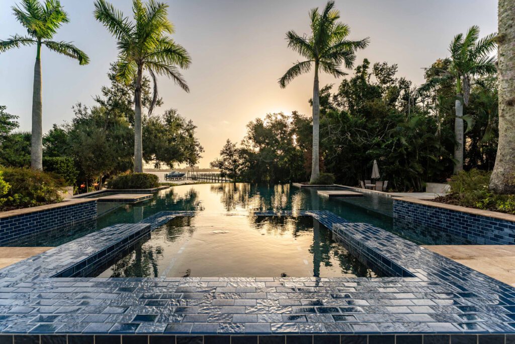 Luxurious pool with blue tile accents, surrounded by palm trees and lush greenery, reflecting sunset light, showcasing Florida Family Pools' custom designs.